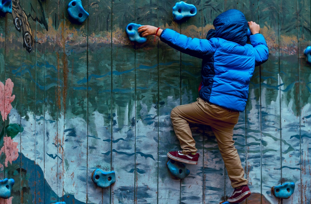 Boy in blue jacket reaching for rock climbing hold 