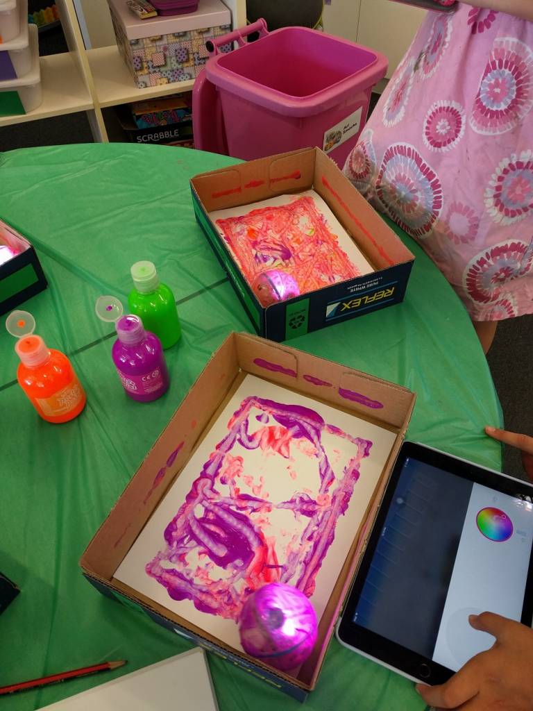 Sphero Art activity. Sphero robots painting canvas inside boxes. Paints and tablet on table.
